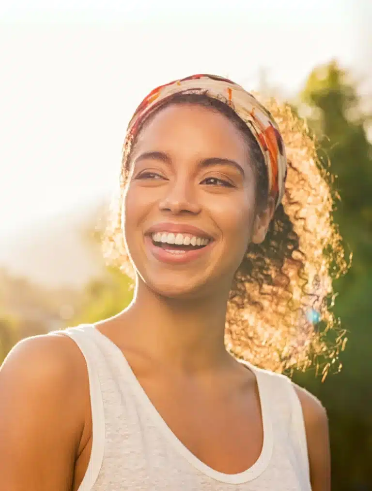 Smiling woman outdoors in sunlight, wearing a headband and white tank top.