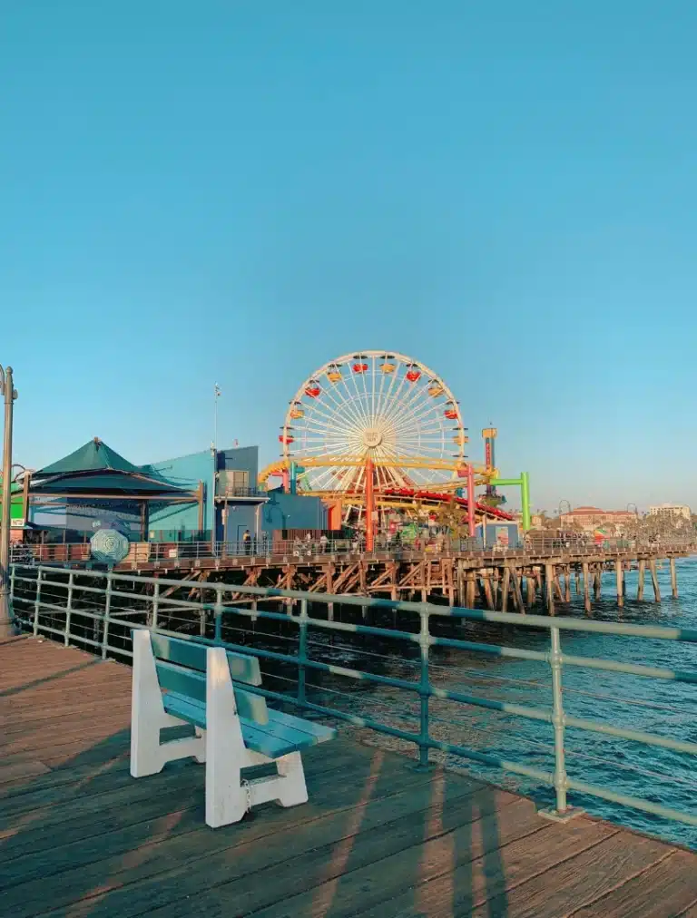 Santa Monica Pier with its iconic Ferris wheel and roller coaster under a bright blue sky, featuring colorful rides, amusement park structures, and American and California flags flying above."