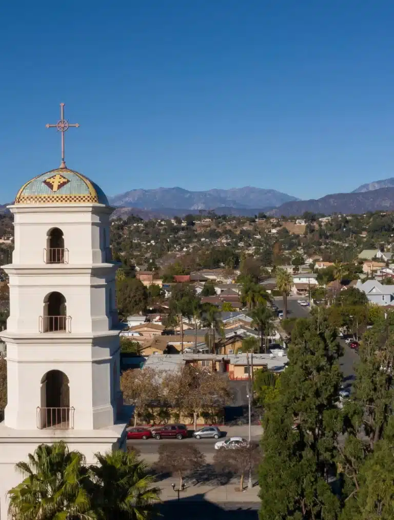 Aerial view of Pomona, California featuring downtown buildings, tree-lined streets, and surrounding mountains under bright daylight.